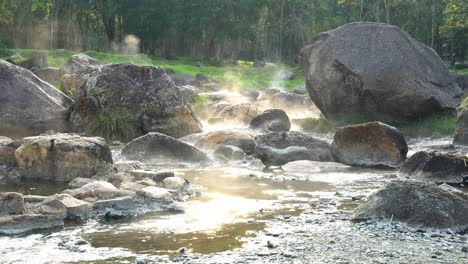 4k slow zoom out of natural hot springs flow along the rocks under evening sunlight, the steam floating up with green grass and forest background. chaeson national park, lampang,thailand