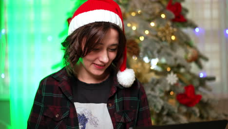 good-looking happy caucasian lady working at laptop. brunette wearing santa cap communicating online. christmas tree at backdrop.