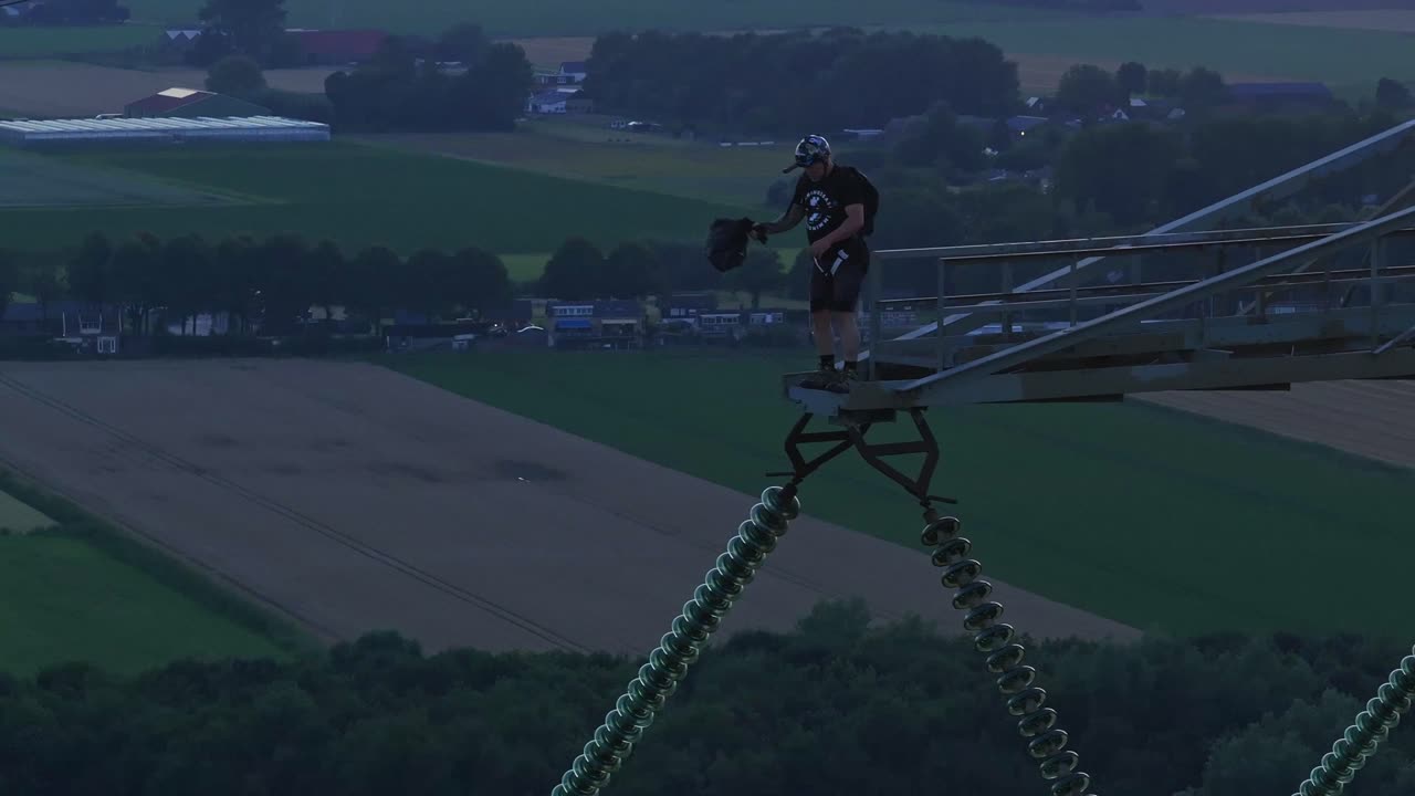 Man standing on a tall structure, preparing to base jump over scenic farmland during sunset