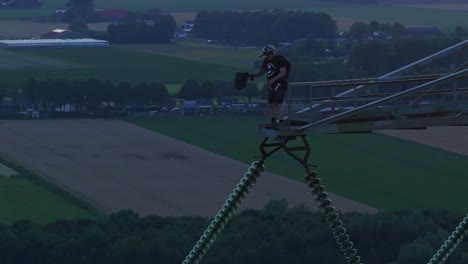 man standing on a tall structure, preparing to base jump over scenic farmland during sunset