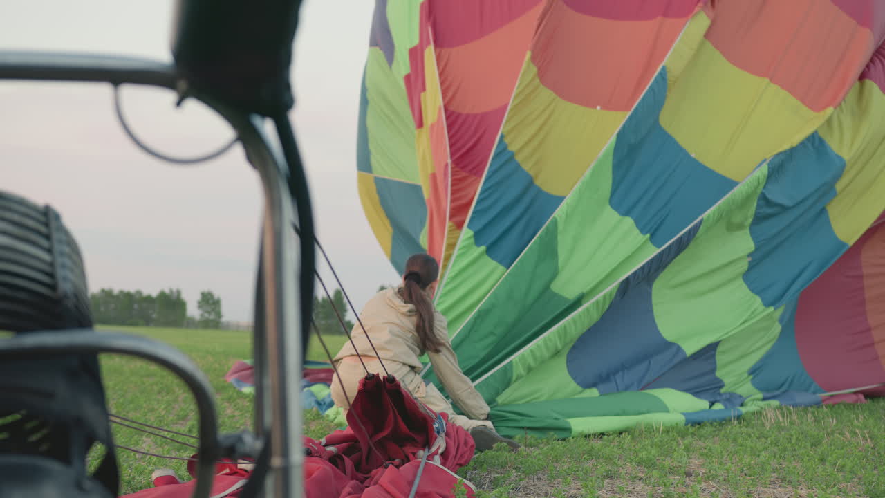 back view of woman in beige jumpsuit organizing collapsed vibrant hot air balloon fabric on green field near metal burner frame during calm evening after flight