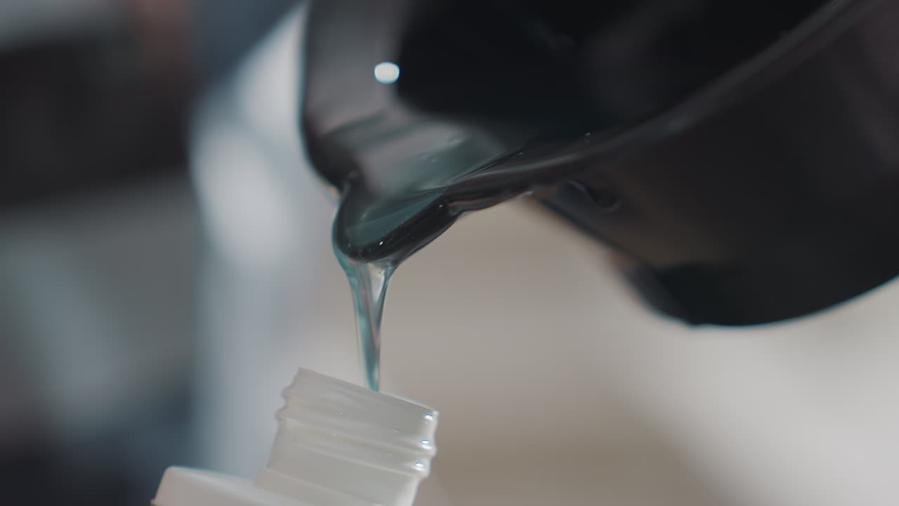 Primer plano de tinte viscoso vertiéndose de un cuenco para mezclar en un pequeño frasco aplicador, goteo cuidadoso y llenado preciso, fondo de estudio tenue y manos concentradas preparando el producto para su aplicación