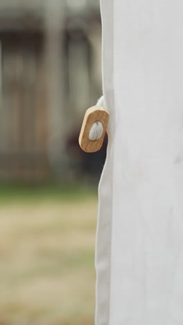 white tent stands in courtyard of medieval settlement. wind shakes dense fabric. people in ancient clothes walk near wooden houses in village on blurred background