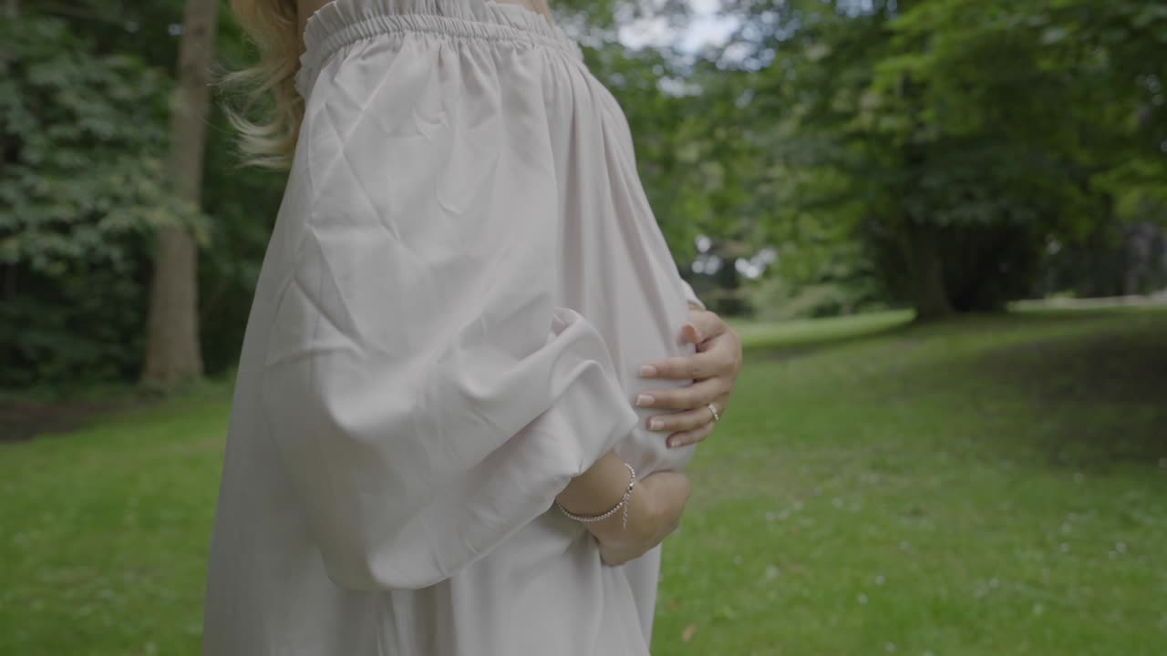 美しい白いドレスを着て、大きなお腹を抱えて緑豊かな庭園を歩く妊娠中の若い女性のクローズアップショット