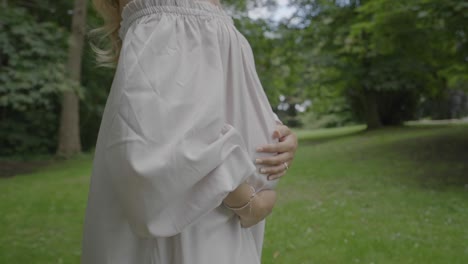 무성한 녹색 정원을 걷는 동안 아름다운 하얀 드레스를 입고 큰 배를 안고 있는 임신한 젊은 여성의 클로즈업 샷