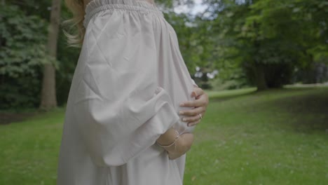 closeup shot of a pregnant young woman wearing a beautiful white dress and holding her large belly, while walking through her lush green garden