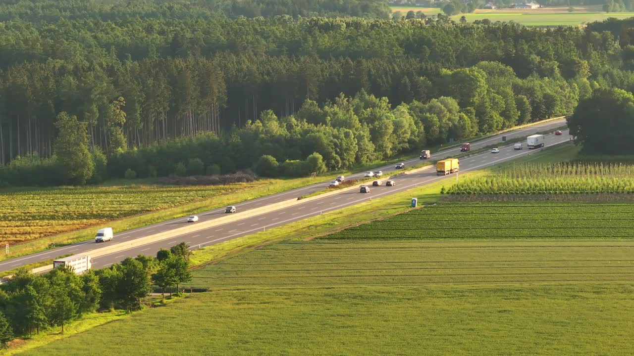 Cars and trucks drive on Germany's A8 highway, surrounded by green fields and forests at sunset