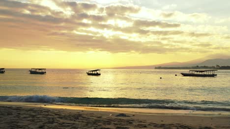 Golden-sunset-with-glowing-yellow-sky-and-grey-clouds-reflecting-on-calm-sea-with-boats-floating-near-beach-in-Indonesia