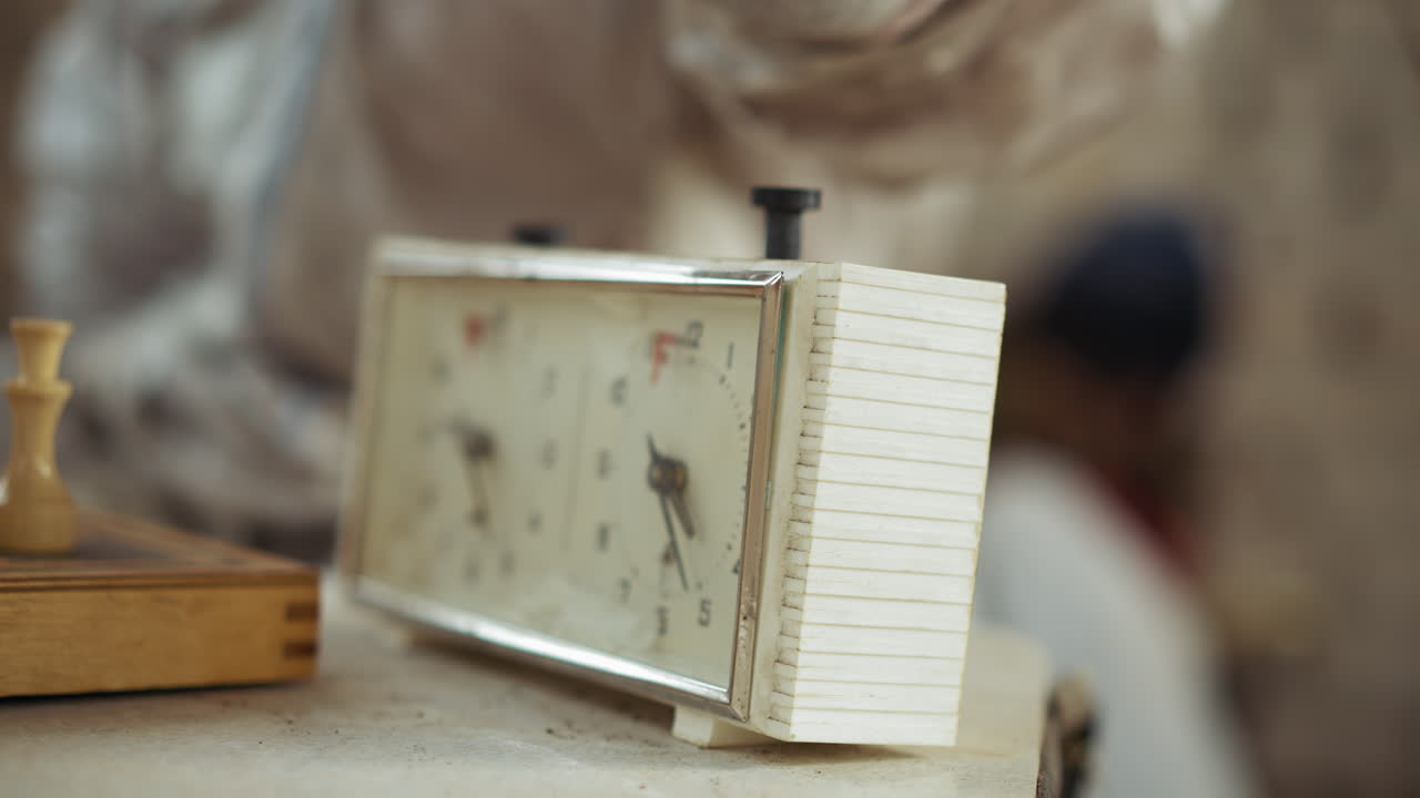 Close up of dark skinned dirty hand in white sleeve pressing mechanical chess clock during indoor match, showing dusty surface and worn timer, with wooden chessboard partially visible beside device