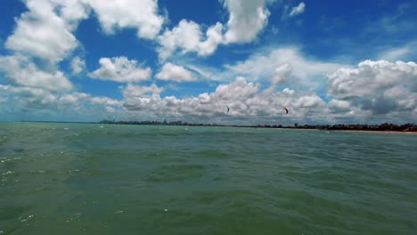 Zwei-Kitesurfer-Surfen-Auf-Dem-Wasser-Mit-Großen-Wolkenkratzern-Der-Tropischen-Stadt-Joao-Pessoa,-Die-Sich-An-Einem-Warmen,-Sonnigen-Sommertag-Mit-Türkisfarbenem-Wasser-Und-Blauem-Himmel-Im-Hintergrund-Abzeichnen