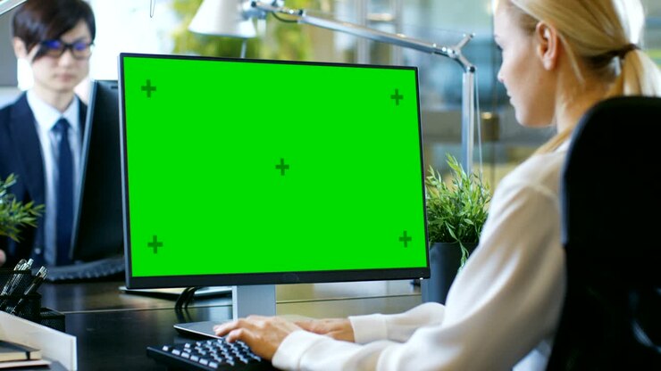 In ufficio, una donna d'affari lavora alla sua scrivania su un computer personale con mock-up a schermo verde. Un collega le siede di fronte. Stanza dall'aspetto moderno con stile vetro e cemento.