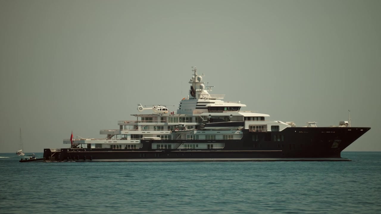 Grande superyacht scuro che naviga in mare aperto sotto un cielo sereno, con un elicottero ben visibile sul ponte.