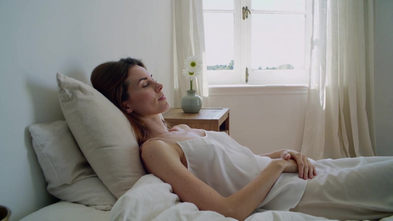 Luce solare che filtra dalla finestra, illuminando una donna sdraiata con un top chiaro che riposa sul letto, con un vaso sul comodino.