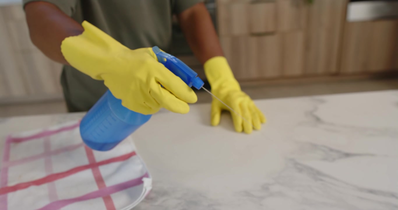 Mujer adulta con guantes amarillos rociando un pulverizador azul sobre una encimera de mármol mientras limpia la cocina.