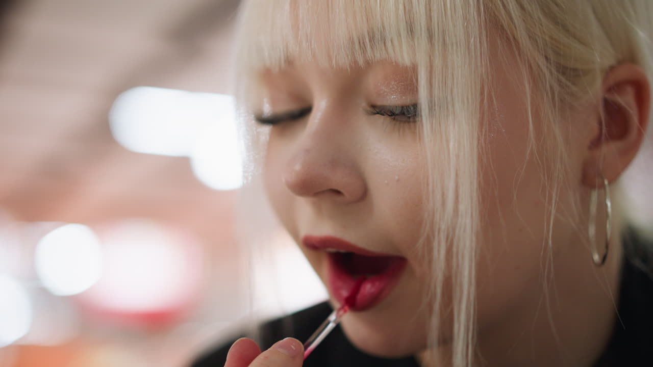 Close up of blonde woman with bangs and hoop earrings carefully painting lips with focused expression, holding lipstick applicator, showcasing beauty with blurred indoor background