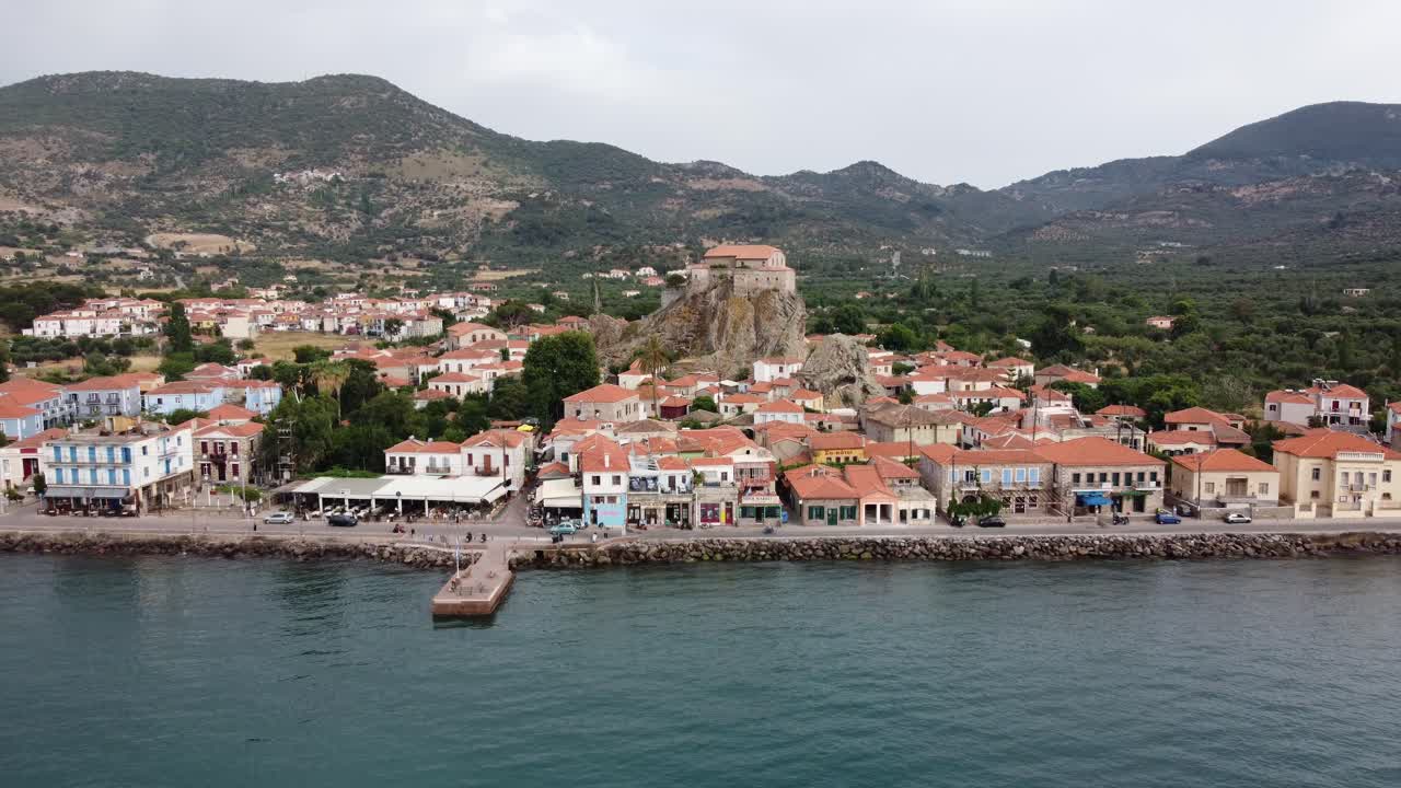 Beachfront Petra village busy street view with traditional houses, shops, tavernas, Virgin Mary Greek Orthodox Church at top of rock in center surrounding mountain landscape, Lesvos, Drone view