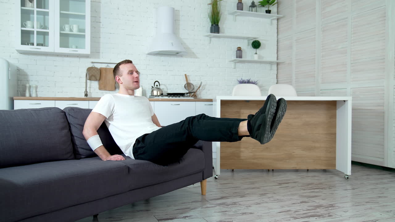 Young man doing sport at the apartment. Guy in white t-shirt, black pants and trainers exercise while sitting on couch. Young fellow doing workout and smiling.