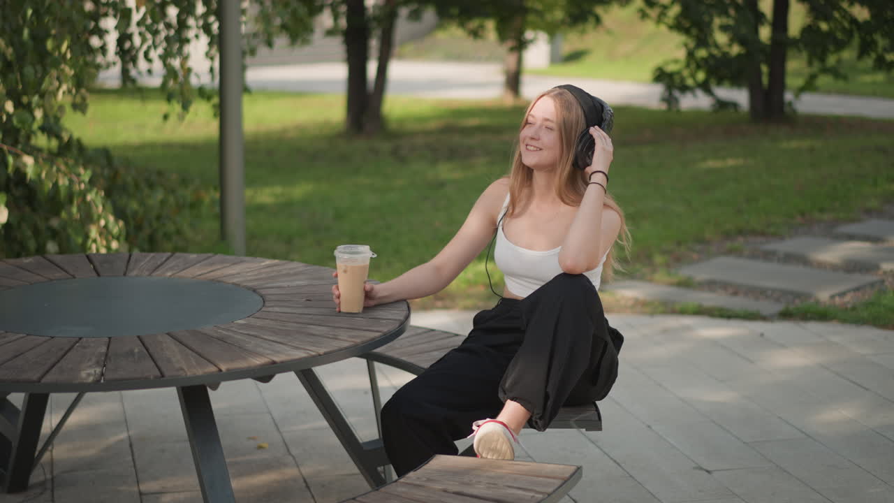 En kvinde sidder ved et bord i parken og nyder sin kaffe, mens hun lytter til musik i sine høretelefoner og vugger i takt til rytmen. Sollyset skinner gennem trækronerne og skaber en pletvis skygge på træbordet. Hun er klædt i et afslappet sommeroutfit, har en latte i hånden og en afslappet holdning. Et ærligt øjeblik af hendes livsstil.