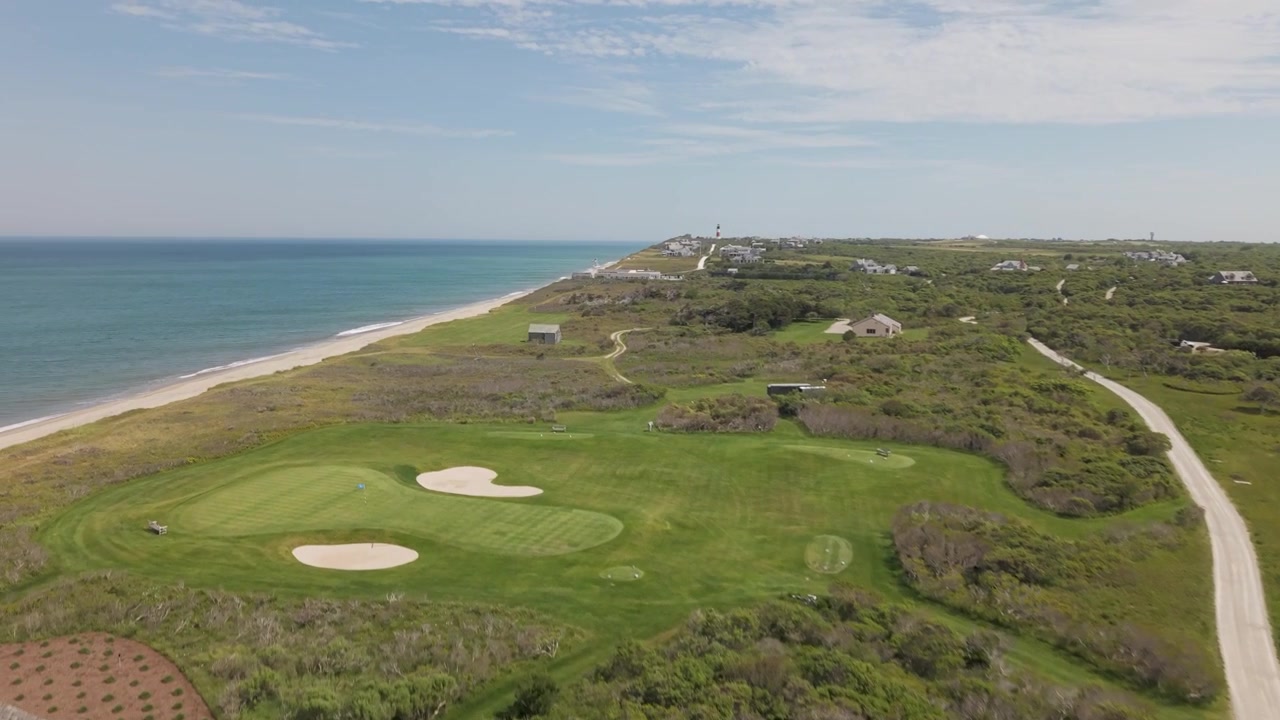 Casa sulla spiaggia con scandole consumate e alto camino in mattoni si affaccia su un vialetto di ghiaia a Nantucket; il lontano faro di Sankaty Head segna la macchia costiera, Massachusetts, USA. Vista aerea con drone