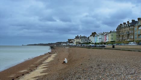 Winter-beach-scene-on-an-English-South-Coast-beach,-of-a-young-woman-sitting-all-alone,-gazing-out-to-sea-looking-pensive-and-sad,-under-a-dark-moody-sky