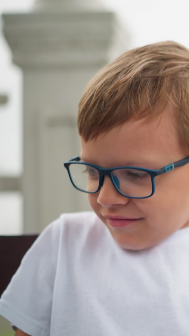 un niño pequeño con gafas se sienta solo con una expresión seria, pareciendo contener una sonrisa mientras se ajusta en la silla