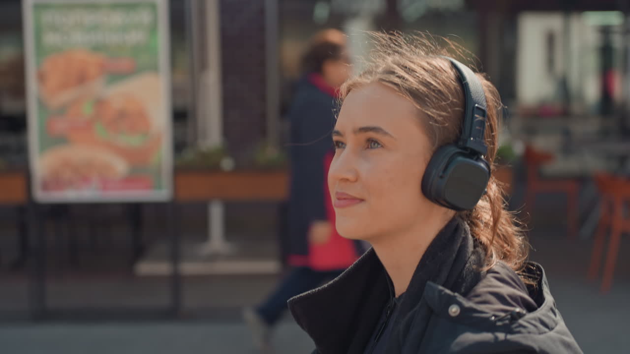 Ung kvinne med hodetelefoner som spaserer i bygaten forbi en utendørs kafé, sollys i håret, over-ear headset, svart kåpe, avslappet smil, ærlig pendlerøyeblikk, dempet urbant lydlandskap