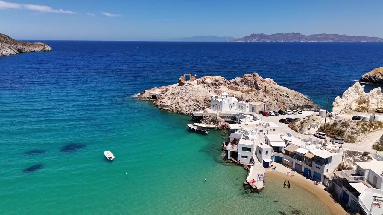 La spiaggia di Firopotamos e le case bianche dei pescatori si rivelano in una veduta aerea su Milos, con una piccola chiesa su un promontorio roccioso, auto parcheggiate, due barche e persone accanto a acque basse turchesi