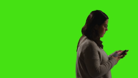 woman walking across frame looking at text message or browsing online on mobile phone against green screen background with low key lighting