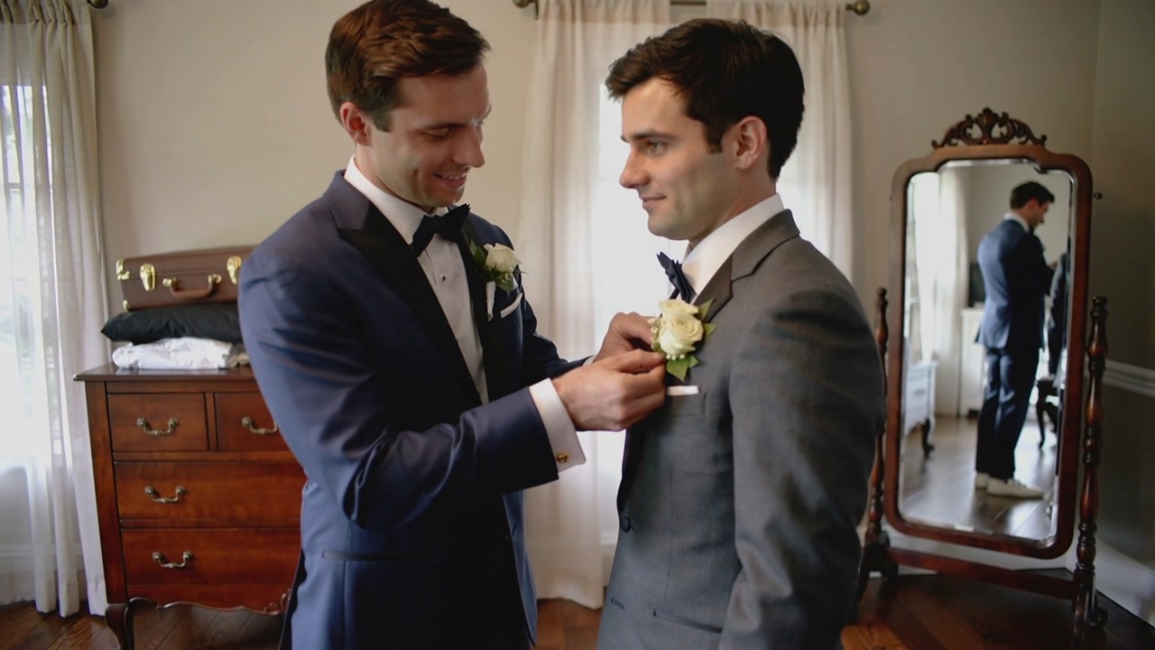 Pinning navy-suited man fastening white boutonniere on grey suit preparing for wedding, with mirror