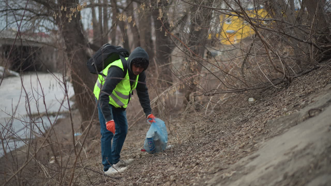 Volunteer in reflective vest and hoodie picking up litter with gloved hands, placing waste into blue plastic bag during environmental cleanup on dry riverbank with bare trees and scattered debris