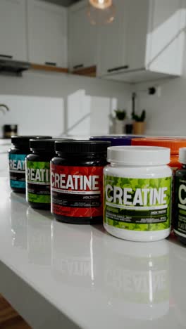 multiple jars of creatine supplements, in various colors and sizes, sit on a white kitchen counter, reflecting in its glossy surface, with white cabinets and a small plant in the background