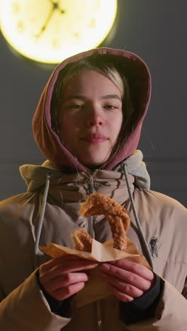 mujer joven de pie frente a un reloj con pastel en la mano, disfrutando de su bocadillo al aire libre por la noche, entorno urbano, ambiente acogedor, y reloj iluminado en el fondo