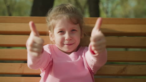 smiling little girl shows thumbs-up on bench in spring park