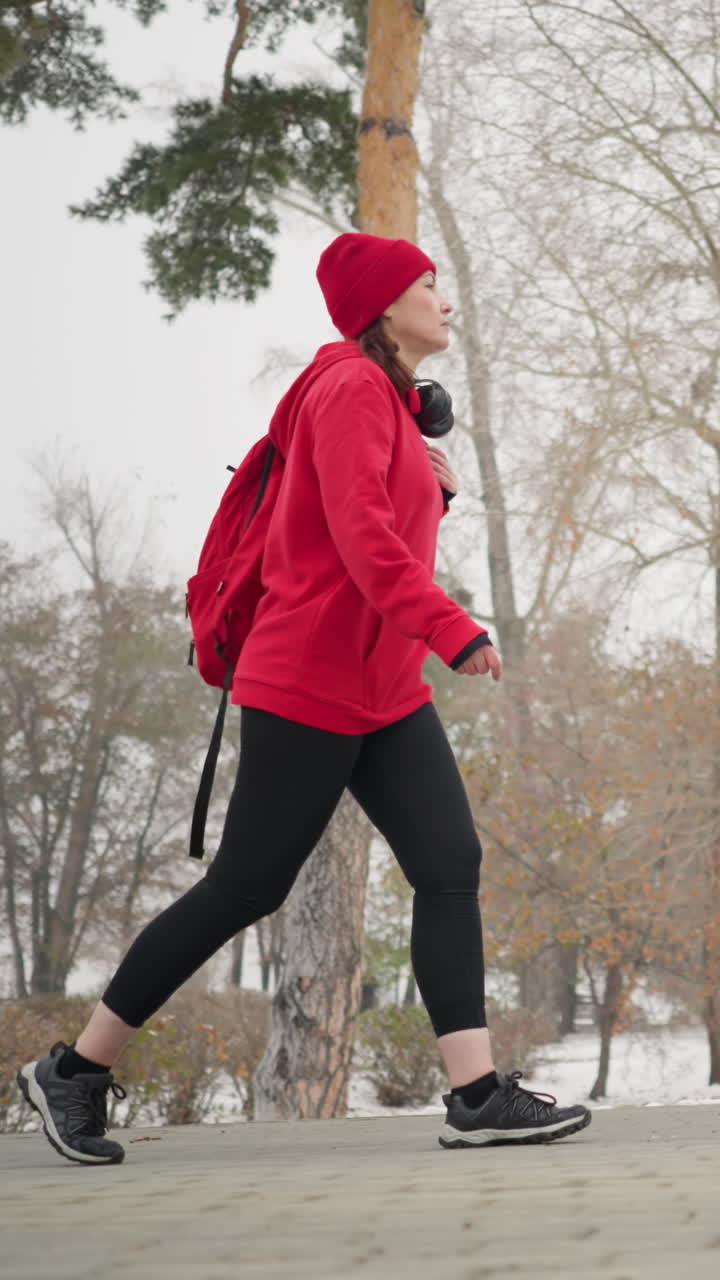 Lower angle view of freelancer with bag over shoulder walking along interlocked pathway surrounded by snowy park scenery, colorful bushes, and tall trees in foggy atmosphere during winter