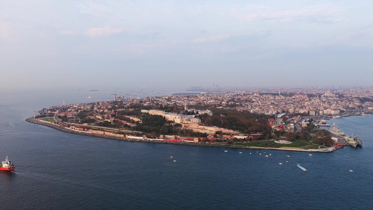 Vídeo impressionante de drone da Península de Hel revela uma cidade costeira extensa, vegetação exuberante, navio de carga vermelho e pequena embarcação perto da costa. A luz suave e nebulosa do dia banha delicadamente os edifícios densos abaixo