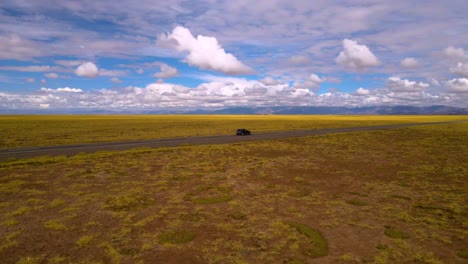 Disparo-De-Un-Dron-Volando-Alrededor-De-Un-Automóvil-Gris-Conduciendo-Por-Una-Carretera-Recta-A-Través-Del-Lago-Salado-De-Salinas-Grandes-En-La-Frontera-De-Salta-Y-Jujuy,-Argentina