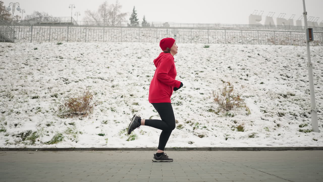 Side view of adult jogging outdoors on interlocked pavement near snowy hillside in winter, runner in red hoodie showcases active lifestyle, with bare trees, lamp posts, and serene urban setting