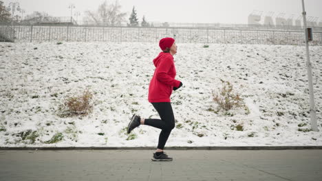 side view of adult jogging outdoors on interlocked pavement near snowy hillside in winter, runner in red hoodie showcases active lifestyle, with bare trees, lamp posts, and serene urban setting