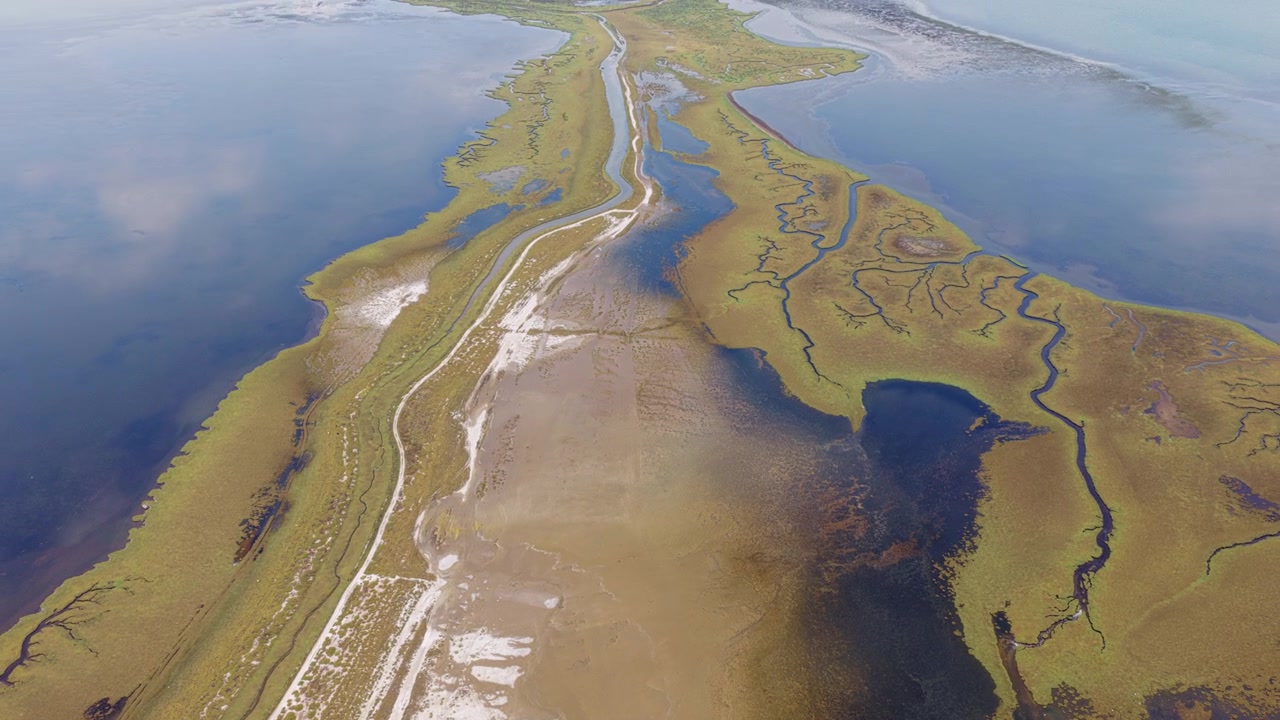 Maestosa prospettiva aerea che sorvola un paesaggio costiero unico con una stretta striscia di terra, intricate saline e acque blu calme che riflettono il cielo in una giornata di sole