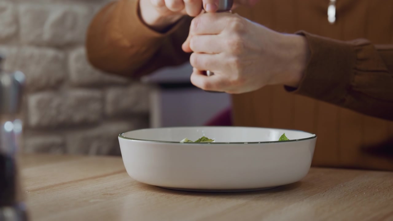 Close up of person seasoning food in white bowl on wooden table. Hands holding pepper grinder sprinkle salt over green leaves with stone wall and kitchen background visible