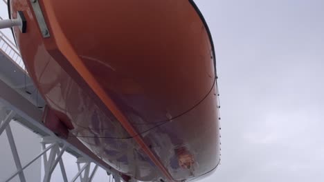 orange lifeboat hanging on offshore vessel, viewed from the bottom against grey cloudy sky