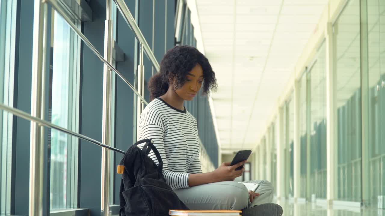 African american woman student sitting on the floor with a phone in the university. New modern fully functional education facility. Concept of online education