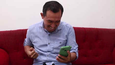 Happy-succeed-asian-man-using-mobile-phone-while-sitting-on-the-sofa-at-home