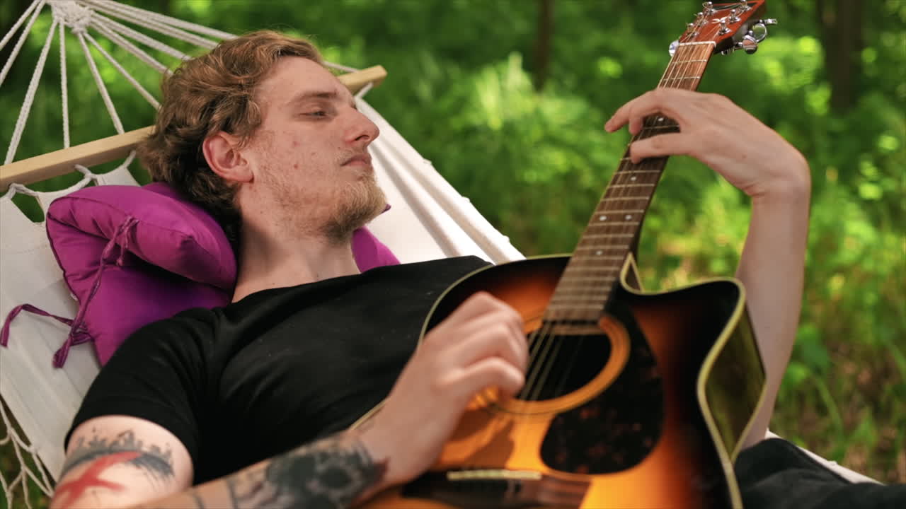 Young man lying on hammock is playing guitar. Greenery around. Glamping. Slow motion
