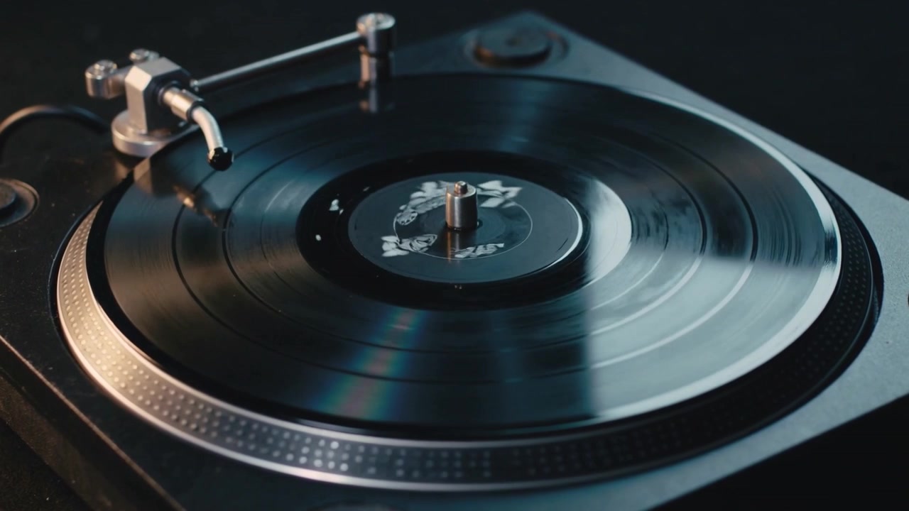 Motore in funzione, disco in vinile che gira in senso orario su un tavolo da studio in penombra, con il braccio del giradischi sospeso.