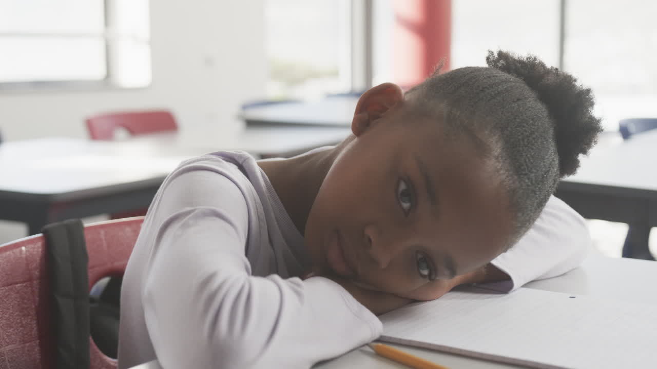 In school, African American girl resting head on desk with notebook and pencil, looking tired