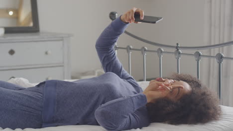 woman wearing pyjamas posing and taking selfies on mobile phone lying on bed