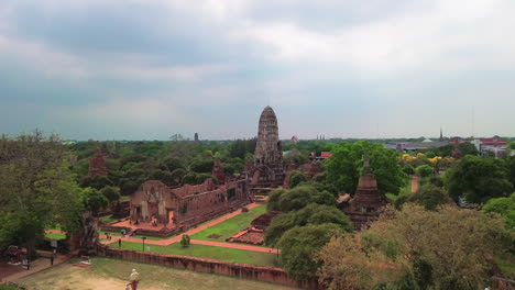 drone gradual approach over temple ruins and green trees of ayutthaya, thailand during calm daylight with cloudy sky