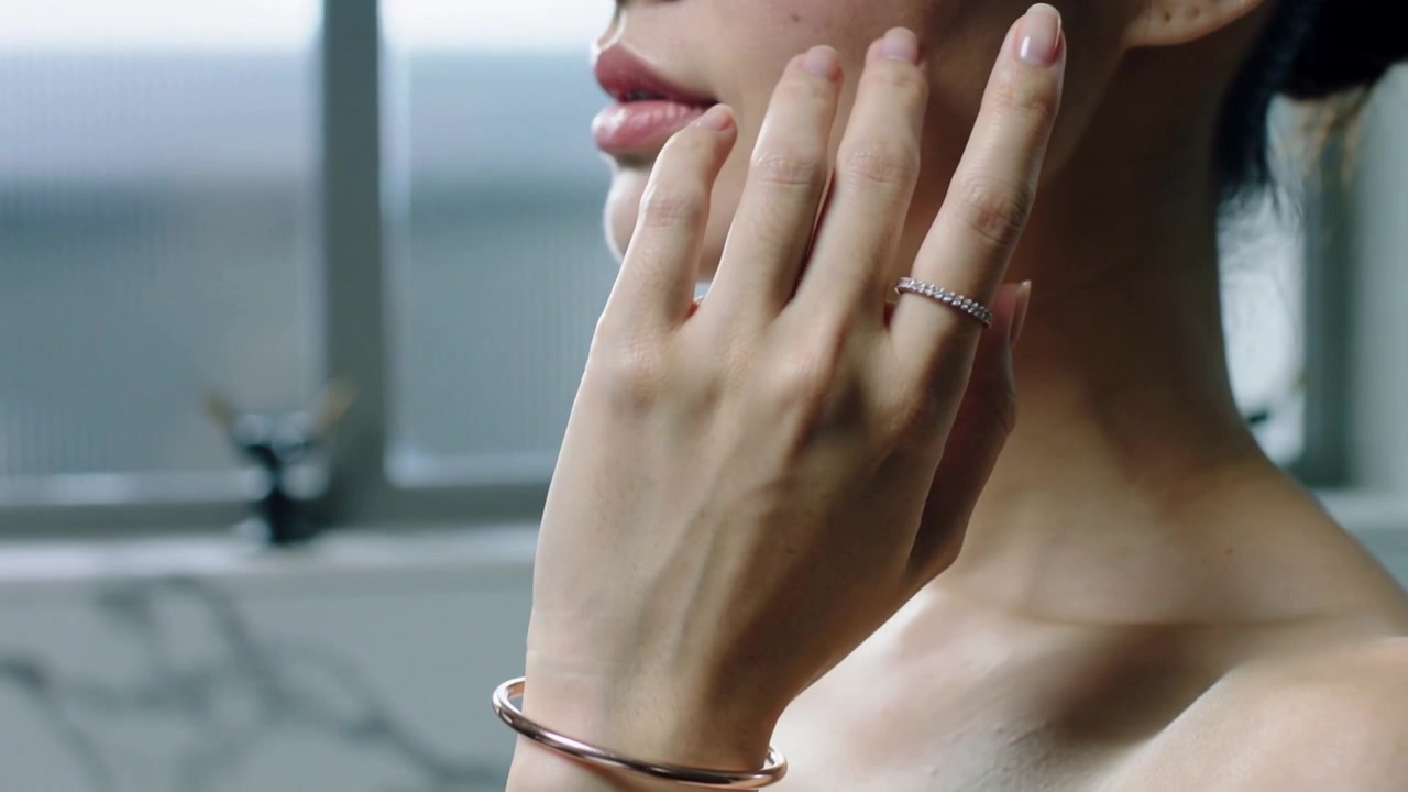 Dopo aver applicato un po' di crema, una donna asiatica senza spalline si strofina lungo la mascella al lavandino, con anello e braccialetto visibili e spazio per il testo.