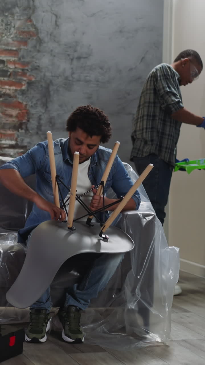 African-American guy paints wall while brother assembles chair in living room. Black friend do domestic repair together. Reconstruction of apartment
